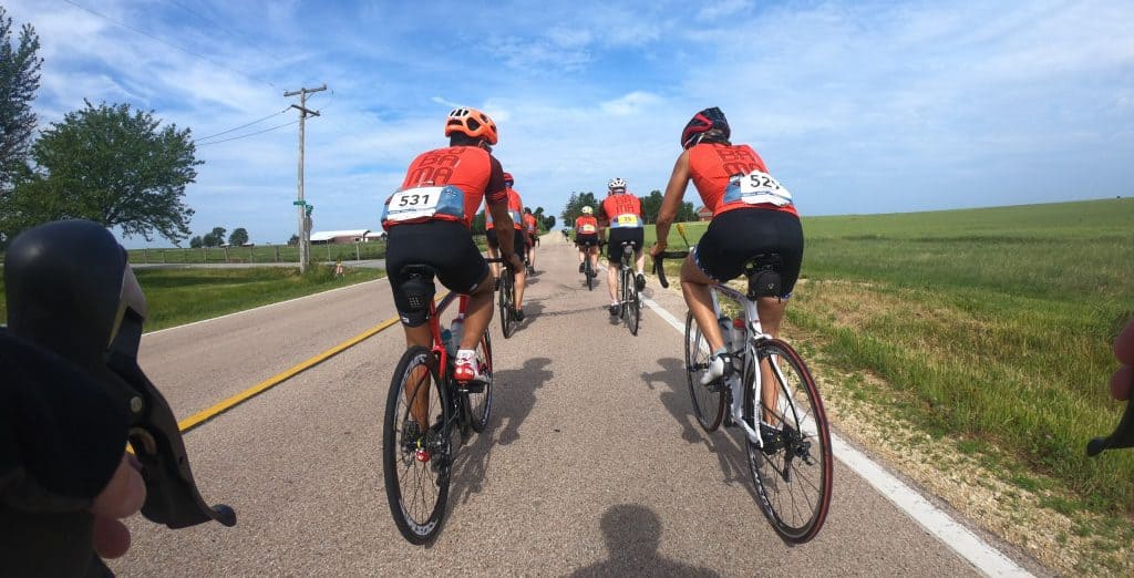 Looking forward on two rows of serious cyclists in red-orange jerseys and helmets on road bikes down a country two-lane road.