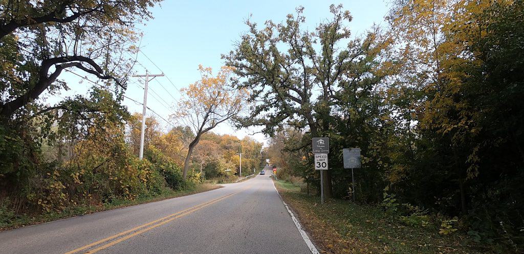 Looking down a two-lane, tree-lined road under a blue sky, with a Welcome to Long Grove sign and Speed Limit 30 sign ahead on the right.