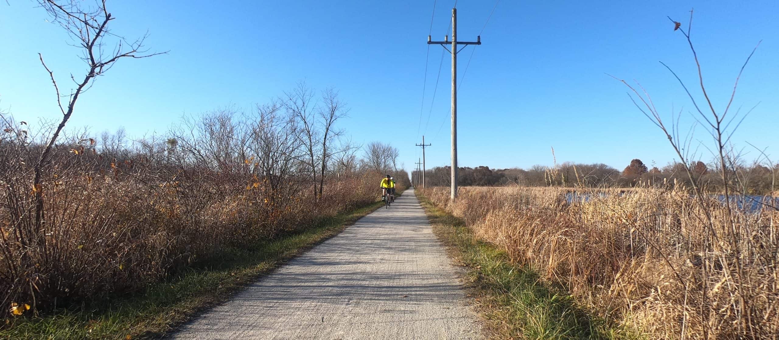 Looking down a gravel trail through prairie, with two bicyclists in the distance, one in a bright yellow jacket, and an electric pole on the right.