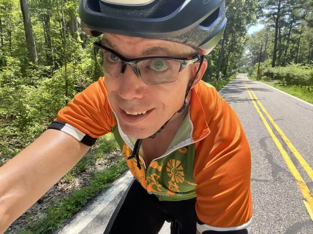 A cyclist in helmet and slightly shaded eyeglasses looks head-on into a selfie with a road through the forest behind him.