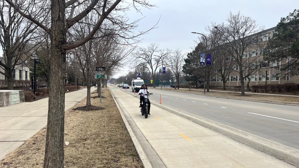 A cyclist riding on a suburban sidewalk in Chicago during winter, with trees and buildings in the ba.