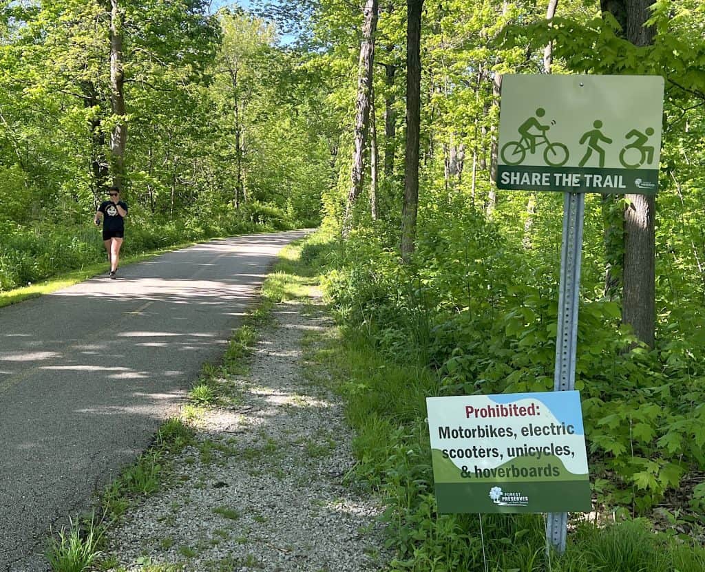 A person walking on a shaded trail with a "Share the Trail" sign for cyclists and pedestrians, in a.